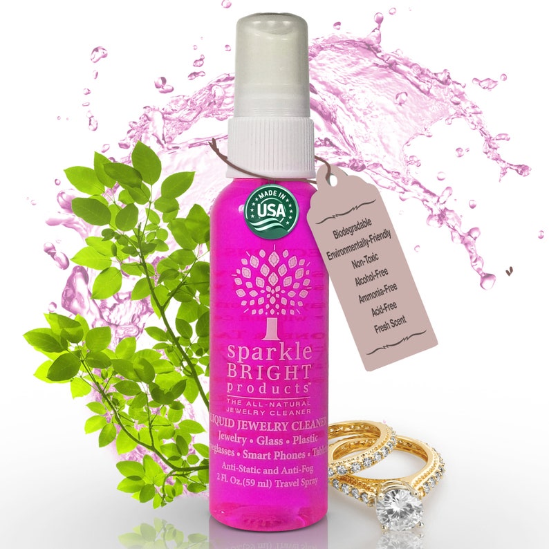 Puede incluir: Botella rosa de limpiador de joyas totalmente natural Sparkle Bright Products con un pulverizador blanco. La botella est&aacute; etiquetada con el texto "Limpiador de joyas l&iacute;quido" y "1 fl. oz. (59 ml) Spray de viaje". La botella est&aacute; rodeada de gotas de agua rosa y hojas verdes.