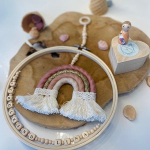 May include: A pink, gold, and white rainbow macrame hanging with wooden beads spelling the name "Lina" and "Schulkind".