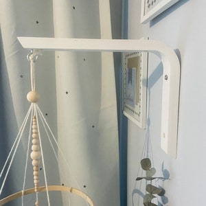 May include: White wooden baby mobile arm mounted on a wall with a white wooden mobile hanging from it. The mobile has a wooden ring with white yarn and wooden beads.