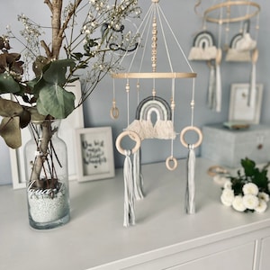 May include: A wooden baby mobile with a rainbow, clouds, and wooden rings. The mobile is hanging from a white ceiling and has a beaded chain with the name "Elias" spelled out.