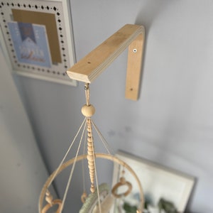 May include: A wooden mobile arm attached to a wall with a white mobile hanging from it. The mobile has a wooden ring with wooden beads, feathers, and rings hanging from it.