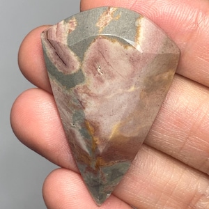 May include: A faceted, shield-shaped gemstone featuring a marbled pattern of muted mauve, dusty rose, earthy brown, and slate gray tones. The stone has a smooth, polished surface with distinct geometric facets along the top edge and a pointed base.