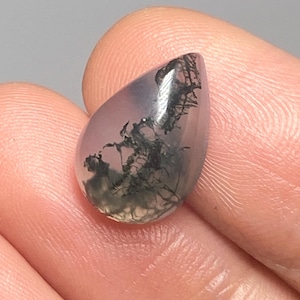 May include: A teardrop-shaped gemstone with a pale lavender tint. The stone has dark green and black inclusions, creating a miniature landscape effect. The polished surface reflects light, showcasing the natural patterns within the stone.