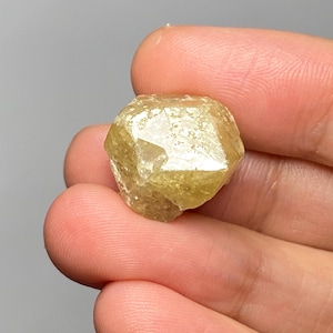 May include: A small, translucent, pale yellow gemstone with a multifaceted, geometric crystal structure. The stone features sharp, angular edges and a polished, reflective surface that catches light, displaying varying shades of light gold and cream.