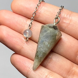 May include: A faceted, cone-shaped labradorite gemstone pendulum suspended from a silver-toned metal chain. The stone displays a greyish-green hue with subtle iridescent flashes. A small, round, translucent bead is attached to the chain.