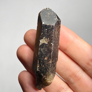 May include: A dark brown, natural crystal with a faceted top and a rough, textured surface. The crystal is held in a hand, showcasing its elongated, prismatic shape and earthy tones. The crystal's color ranges from deep brown to lighter tan.