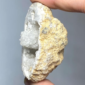 Peut inclure: Une main tient une géode, révélant un intérieur creux rempli de cristaux de quartz clairs et étincelants. L'extérieur est une roche rugueuse de couleur beige avec une surface texturée. La géode est un spécimen géologique naturel.