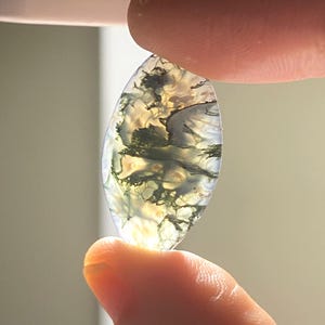 May include: An oval gemstone with a translucent quality, displaying internal patterns of green and black. The stone is held between fingers, showcasing its natural, organic appearance. Light passes through, accentuating the intricate details within.