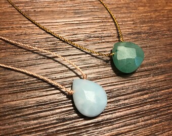 May include: Two necklaces with teardrop-shaped gemstone pendants. The top necklace has a green gemstone pendant on a gold cord. The bottom necklace has a light blue gemstone pendant on a beige cord.