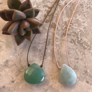 May include: Two necklaces with green gemstone pendants on a light grey background. The necklaces are made with brown and pink cords.