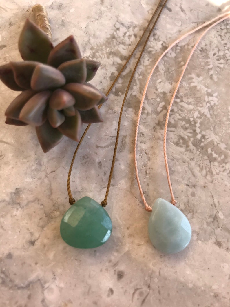 May include: Two necklaces with green gemstone pendants on a light grey background. The necklaces are made with brown and pink cords.