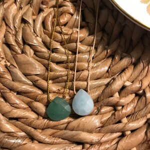 May include: Two teardrop-shaped gemstone pendants on necklaces. The pendant on the left is a dark green colour, and the pendant on the right is a light blue colour. The necklaces are on a woven surface.