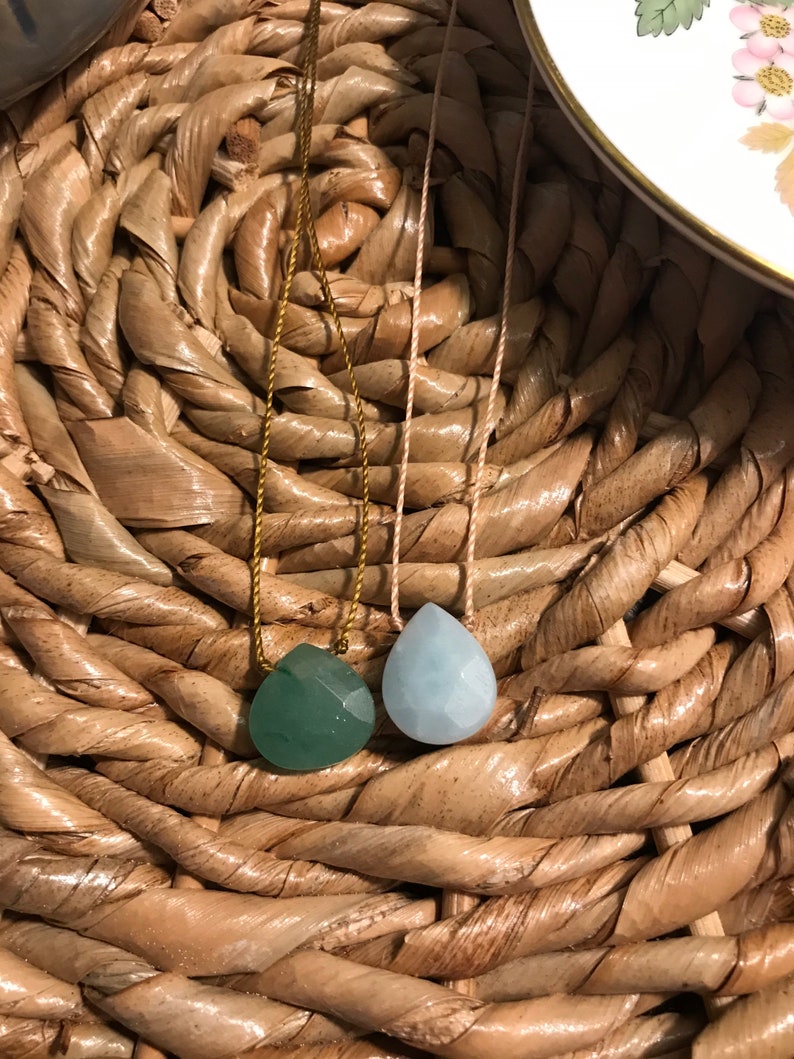 May include: Two teardrop-shaped gemstone pendants on necklaces. The pendant on the left is a dark green colour, and the pendant on the right is a light blue colour. The necklaces are on a woven surface.