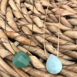 May include: Two necklaces with teardrop-shaped green and blue gemstone pendants on a woven wicker background.