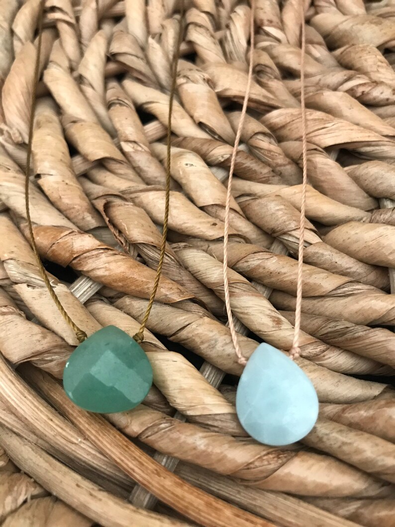 May include: Two necklaces with teardrop-shaped green and blue gemstone pendants on a woven wicker background.