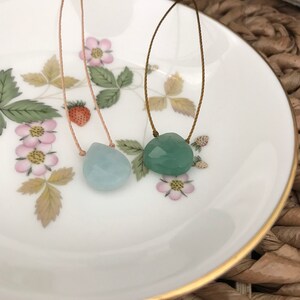 May include: Two necklaces with light blue and green gemstone pendants on a white plate with a gold rim. The necklaces are made with thin brown and beige cords.