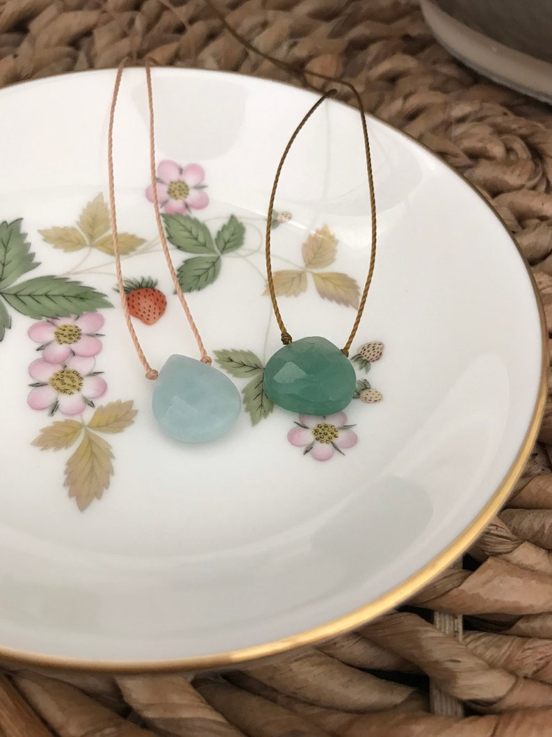 May include: Two necklaces with light blue and green gemstone pendants on a white plate with a gold rim. The necklaces are made with thin brown and beige cords.