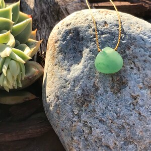 May include: A green sea glass pendant necklace on a gold cord, resting on a grey rock.