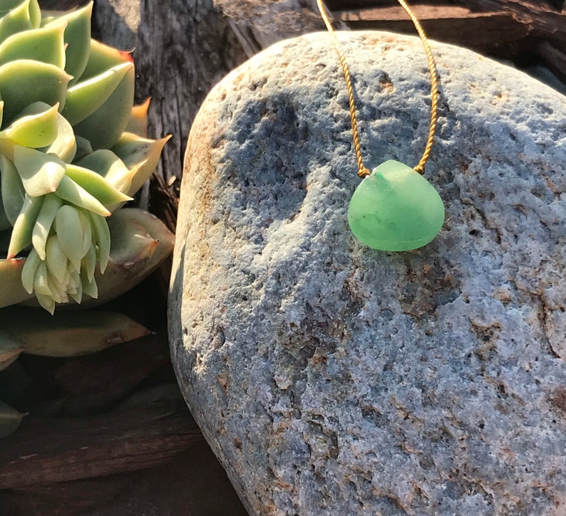 May include: A green sea glass pendant necklace on a gold cord, resting on a grey rock.