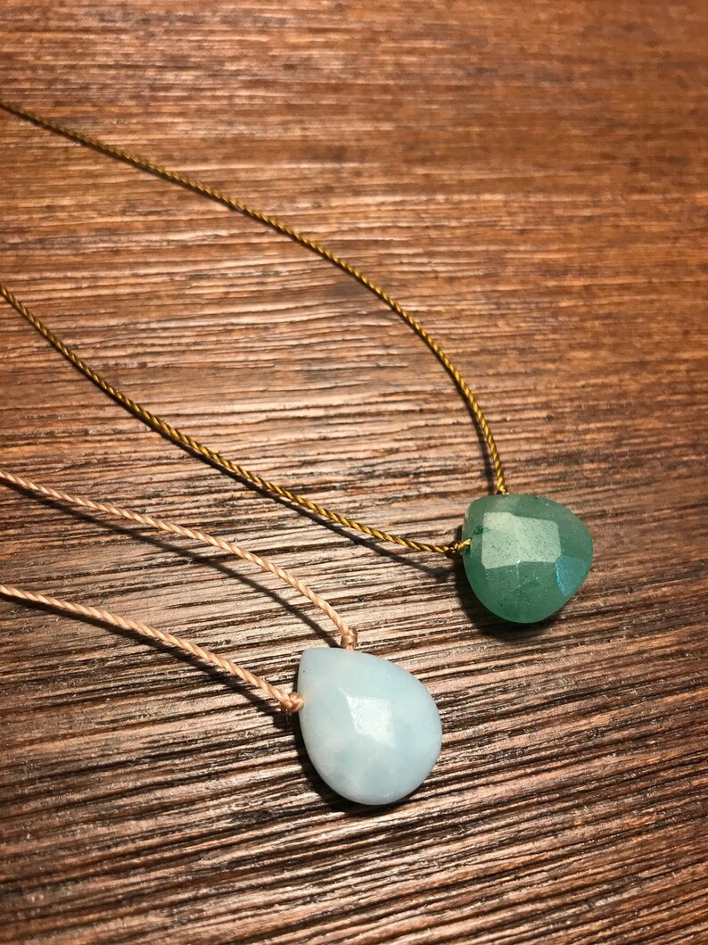 May include: Two necklaces with teardrop-shaped gemstone pendants. The top necklace has a green gemstone pendant on a gold cord. The bottom necklace has a light blue gemstone pendant on a beige cord.