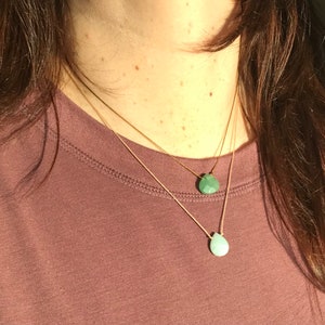 May include: Two delicate gold necklaces with teardrop-shaped green gemstone pendants. The necklaces are layered and worn on a person's neck.