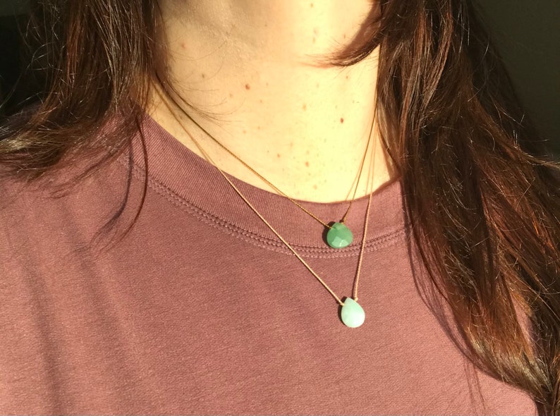 May include: Two delicate gold necklaces with teardrop-shaped green gemstone pendants. The necklaces are layered and worn on a person's neck.
