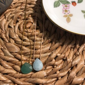 May include: Two teardrop-shaped gemstone pendants on necklaces. The pendant on the left is a dark green colour, and the pendant on the right is a light blue colour. The necklaces are on a woven straw surface.