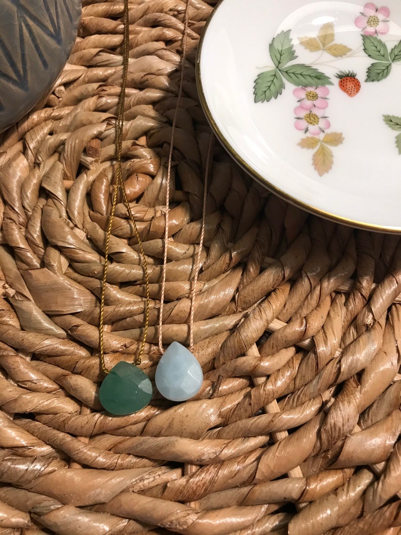 May include: Two teardrop-shaped gemstone pendants on necklaces. The pendant on the left is a dark green colour, and the pendant on the right is a light blue colour. The necklaces are on a woven straw surface.
