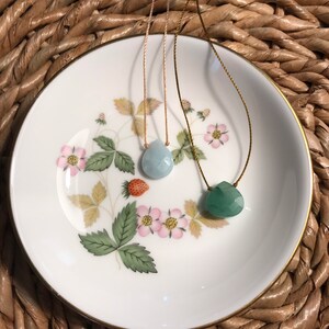 May include: Two necklaces with gemstone pendants on a white plate with floral designs. The pendant on the left is a light blue teardrop shape and the pendant on the right is a green teardrop shape.