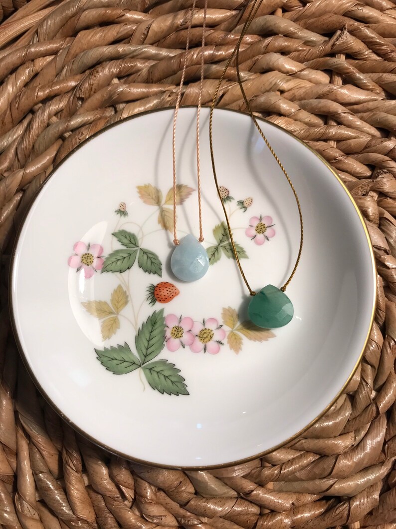 May include: Two necklaces with gemstone pendants on a white plate with floral designs. The pendant on the left is a light blue teardrop shape and the pendant on the right is a green teardrop shape.