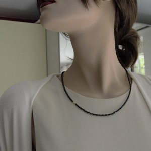 Op de afbeelding: Een zwarte kralenketting met een klein gouden accent. De ketting wordt gedragen door een persoon met donker haar.