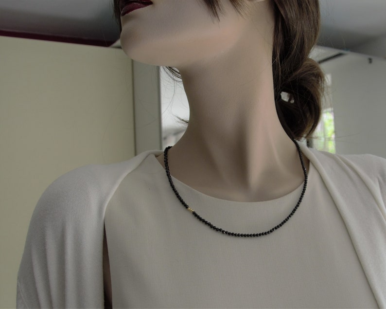 Op de afbeelding: Een zwarte kralenketting met een klein gouden accent. De ketting wordt gedragen door een persoon met donker haar.