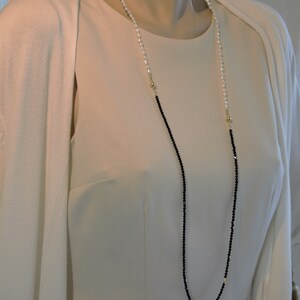 Op de afbeelding: Een lange ketting met zwarte en witte kralen. De ketting is gemaakt van kleine, ronde kralen en heeft een gouden sluiting.