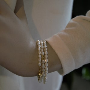 Op de afbeelding: Een set van drie parel armbanden met gouden accenten. De armbanden zijn op elkaar gelegd op iemands pols.
