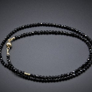 Op de afbeelding: Een zwarte kralenketting met een gouden sluiting. De ketting is gemaakt van kleine, gefacetteerde kralen en is ongeveer 45 centimeter lang.