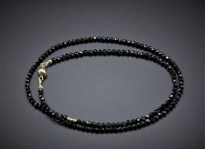 Op de afbeelding: Een zwarte kralenketting met een gouden sluiting. De ketting is gemaakt van kleine, gefacetteerde kralen en is ongeveer 45 centimeter lang.