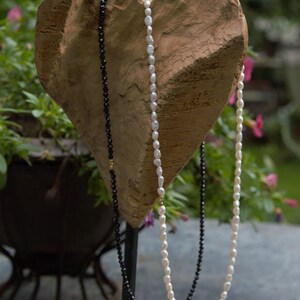 Op de afbeelding: Twee kettingen tentoongesteld op een hartvormige houten standaard. De ene ketting is gemaakt van zwarte kralen en de andere van witte parels.