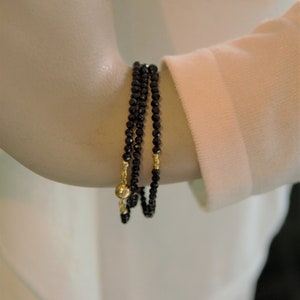 Op de afbeelding: Een drievoudige armband gemaakt van zwarte kralen met gouden accenten. De armband wordt gedragen op de arm van een mannequin.