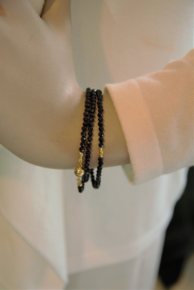 Op de afbeelding: Een drievoudige armband gemaakt van zwarte kralen met gouden accenten. De armband wordt gedragen op de arm van een mannequin.