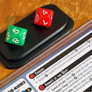 May include: A close-up of a black dice tray holding a red and a green die. The red die shows a zero, and the green die shows a five. A game card is in the background.