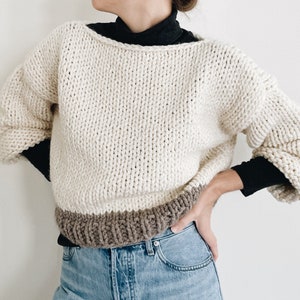 May include: Cream-colored, chunky knit sweater with a wide neckline and long, voluminous sleeves. The bottom hem is a contrasting brown color. The sweater is worn over a black top and paired with light blue jeans. A cozy, handmade garment.