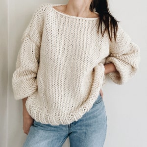 May include: Cream-colored, chunky knit sweater with a wide neckline and long, voluminous sleeves. The sweater has a ribbed hem and cuffs. The style is oversized and comfortable, perfect for casual wear. The sweater is paired with blue jeans.