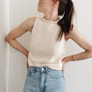 May include: Cream-colored, sleeveless knit top with a high neckline. The top is paired with light blue denim shorts. The outfit is simple and casual, suitable for warm weather. The top is made of a soft, textured yarn.