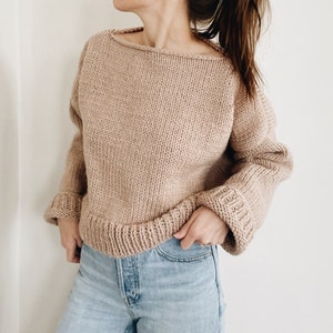 May include: A beige, chunky-knit sweater with a wide neckline and long sleeves. The sleeves have ribbed cuffs, and the bottom hem is also ribbed. The sweater is a neutral color and appears to be made of a soft, warm material.