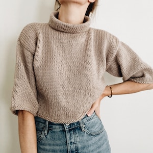 May include: A beige, cropped knitted jumper with a roll neck and short sleeves. The jumper is made of a chunky knit material and is paired with blue denim jeans. A gold bracelet is visible on the arm.