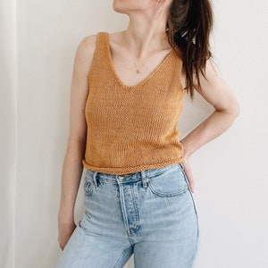 May include: Mustard yellow knitted tank top with a V-neck. The top is sleeveless and has a ribbed hem. The top is paired with light blue denim jeans.