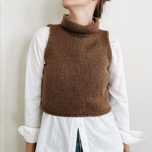 May include: A brown, knitted turtleneck vest. The sleeveless top is made of a thick, textured yarn. The vest is worn over a white button-down shirt and blue jeans. The turtleneck is folded over.
