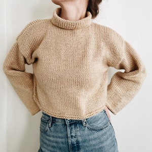 Puede incluir: Un suéter corto de cuello alto beige con mangas largas. El suéter está hecho de un material de punto grueso y tiene un dobladillo y puños acanalados. El modelo lleva vaqueros azules. El suéter es de color neutro y sería adecuado para otoño o invierno.