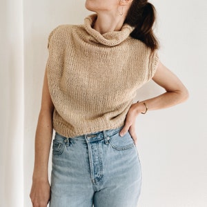 May include: A sleeveless, tan-colored knit top with a turtleneck collar. The top is made of a chunky knit material and has a relaxed fit. The model is wearing light blue jeans.