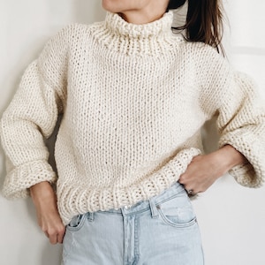 May include: Cream-colored, chunky knit turtleneck sweater. The sweater features a high, ribbed collar, long sleeves with ribbed cuffs, and a ribbed hem. The thick knit creates a textured appearance. The sweater is paired with light wash denim jeans.